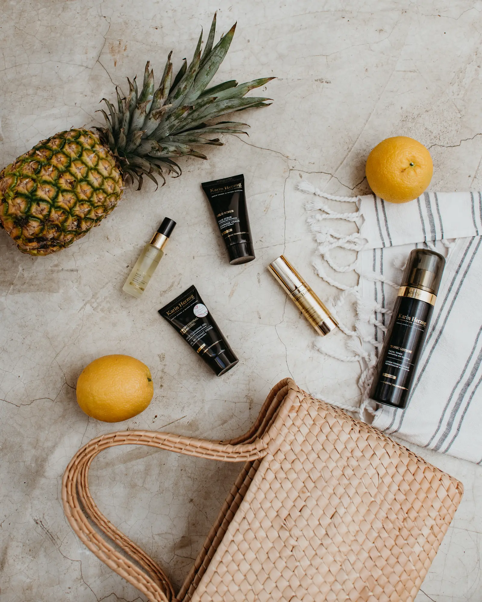 Karin Herzog skincare products on stone, among a pineapple, two oranges, a woven basket and a striped towel